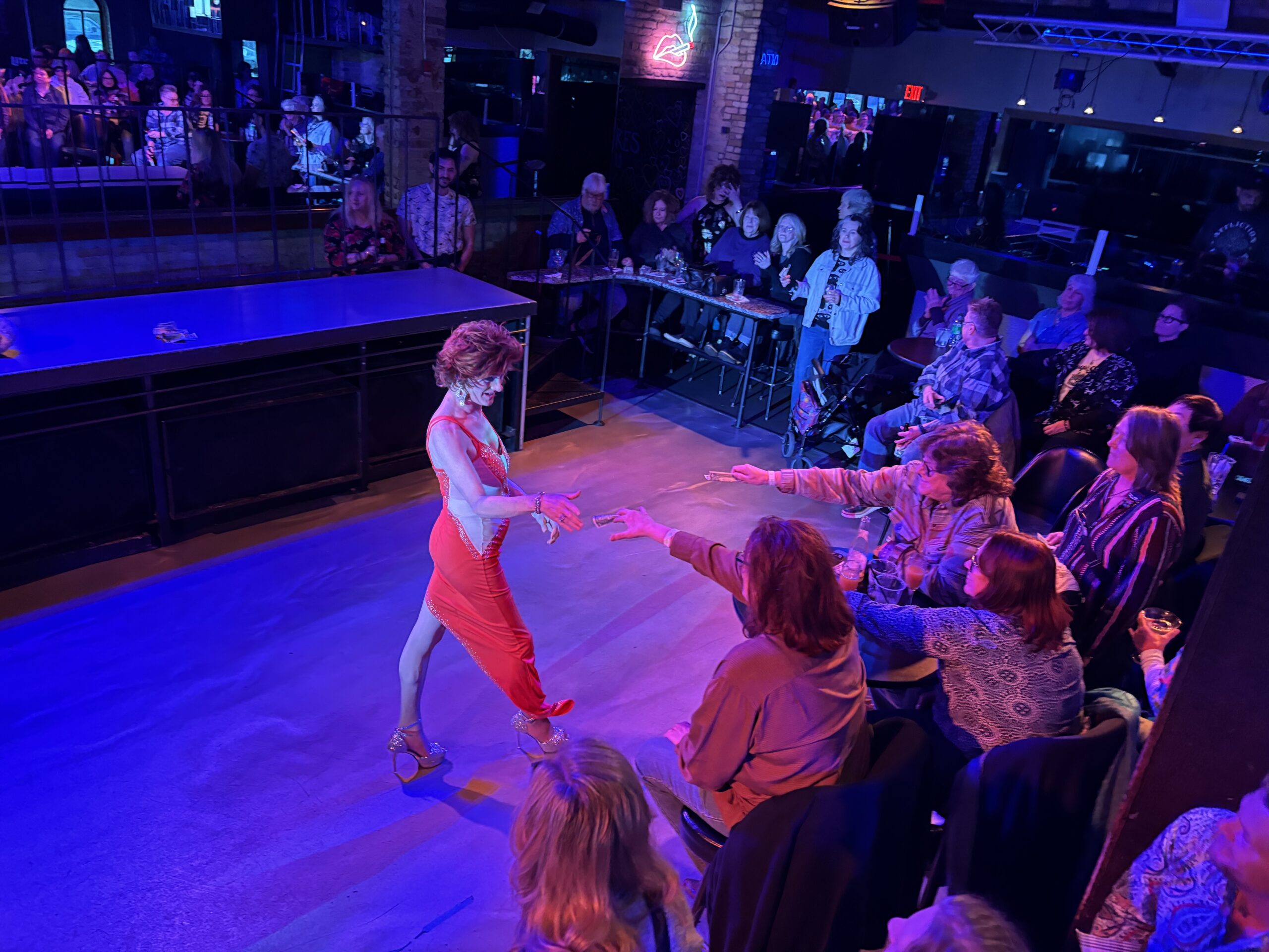 A woman in a red dress dances on a stage in front of an audience, with several people in the front row reaching out toward her.