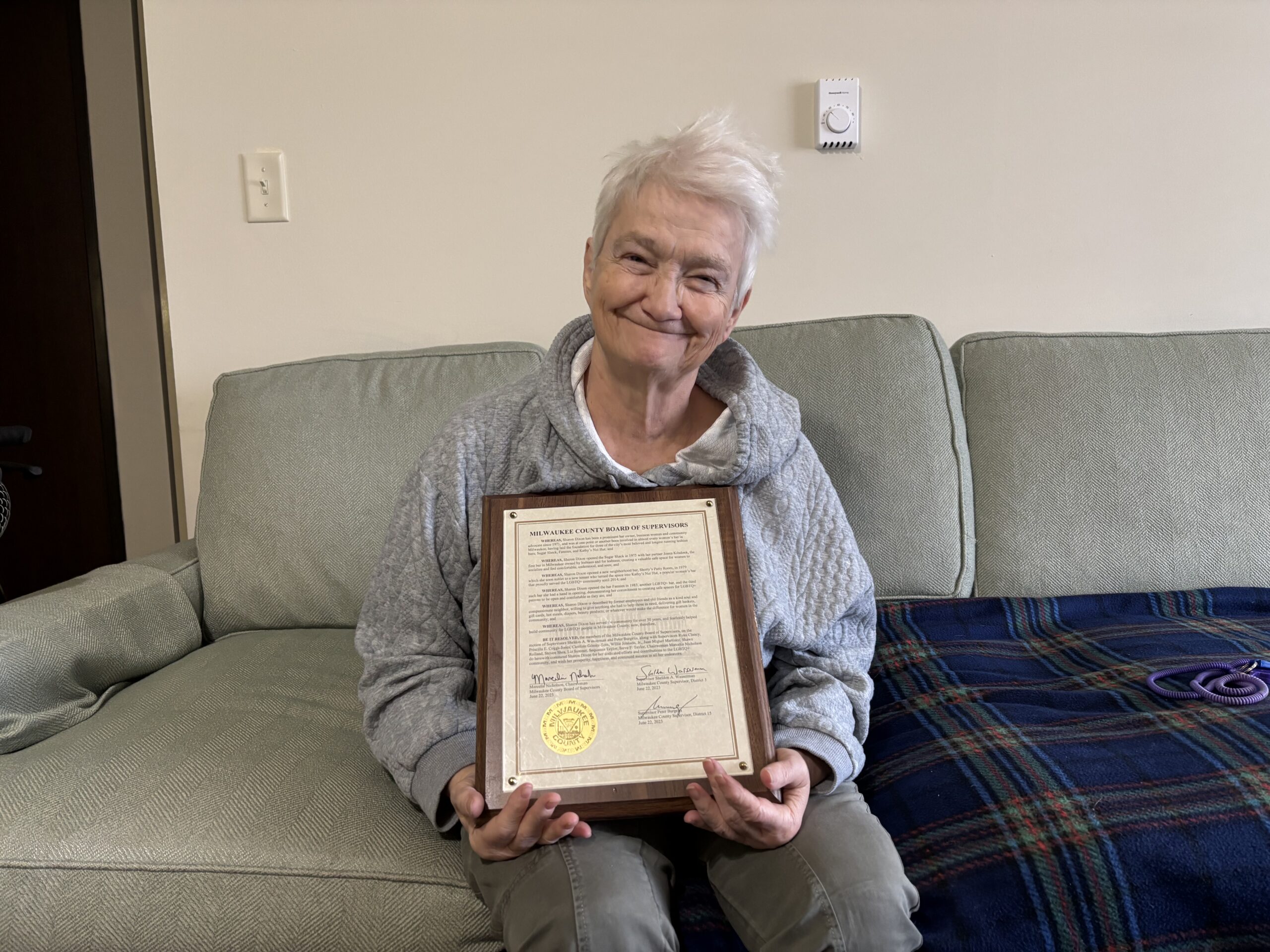 Older person with short white hair smiles while sitting on a couch, holding a framed certificate with a gold seal and signatures.