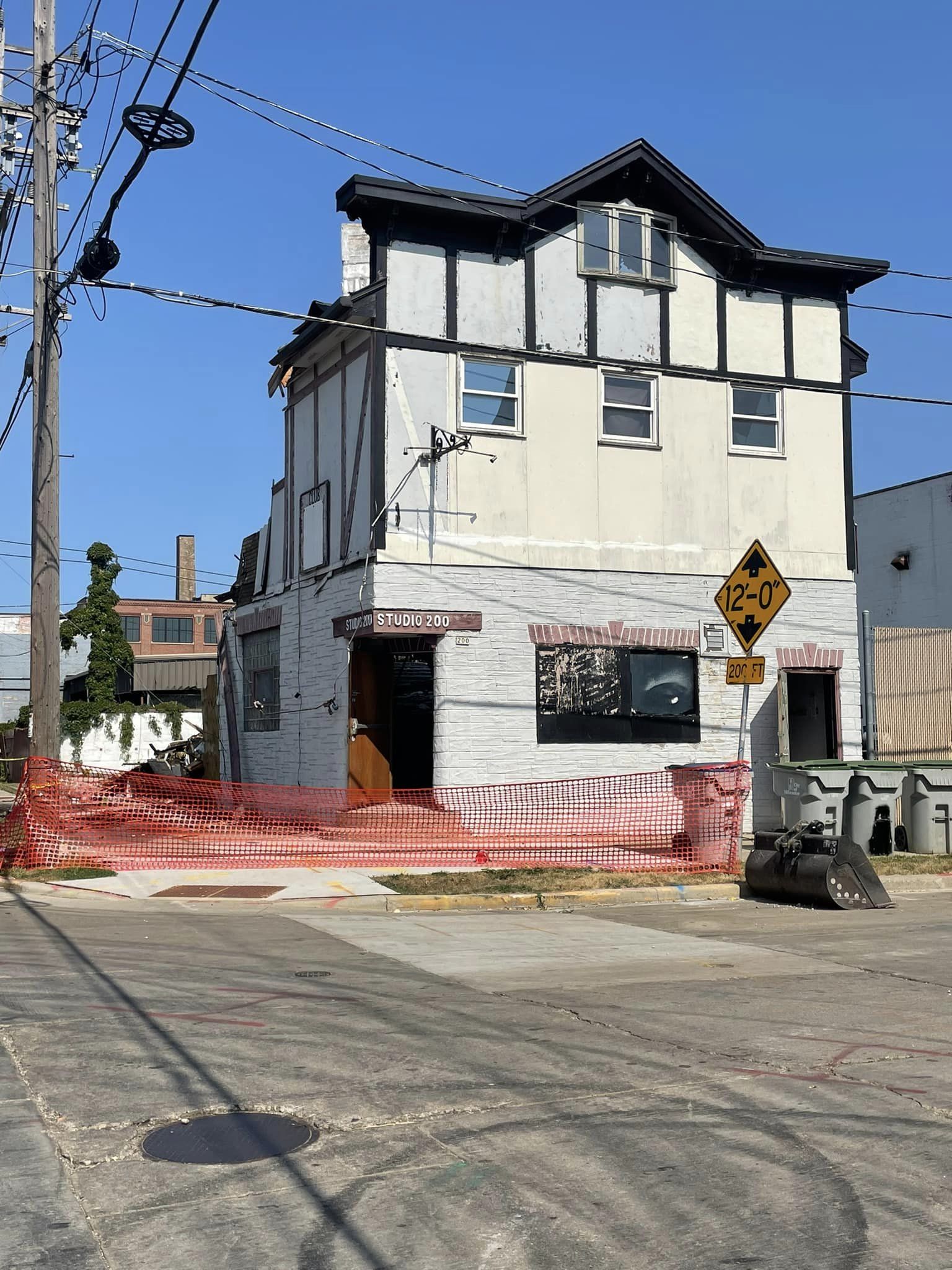 A three-story white building with boarded windows is partially surrounded by orange construction fencing and a 12-0 clearance road sign on a sunny day.